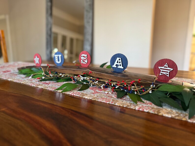 May include: A wooden centrepiece with five round candle holders, each decorated with a red, white and blue patriotic design. The holders spell out "USA" with a star on each end.