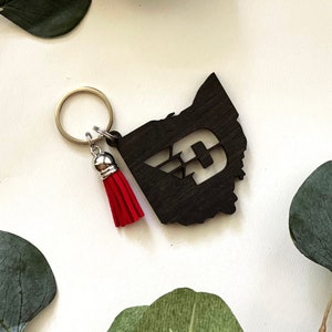 May include: A wooden keychain shaped like the state of Ohio with a black and white letter D inside. The keychain has a silver ring and a red tassel.