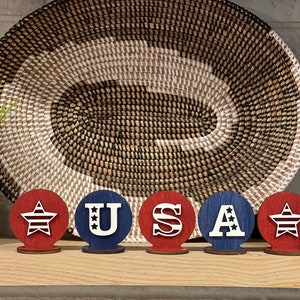 May include: Five wooden circles painted in red, white and blue. Each circle features a different design: a star, the letter U, the letter S, the letter A, and a star. The circles are arranged in a row, spelling out the word "USA".