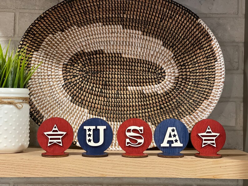 May include: Five wooden circles painted in red, white and blue. Each circle features a different design: a star, the letter U, the letter S, the letter A, and a star. The circles are arranged in a row, spelling out the word "USA".