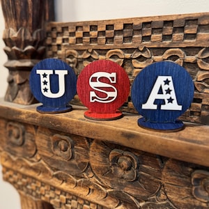 May include: Three wooden circles with the letters "USA" painted in white on a red, white, and blue background. The circles are arranged in a row on a wooden shelf.