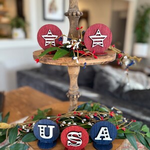 May include: Wooden tiered tray decorated with red, white and blue stars and the letters "USA". The stars and letters are cut out of wood and painted white. The tray is decorated with greenery.