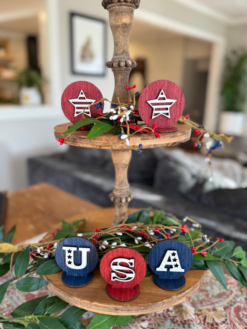 May include: Wooden tiered tray decorated with red, white and blue stars and the letters "USA". The stars and letters are cut out of wood and painted white. The tray is decorated with greenery.