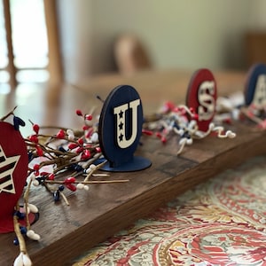 May include: A wooden centrepiece featuring the letters "USA" in red, white and blue. The letters are on individual stands and are surrounded by red berries and white sprigs.