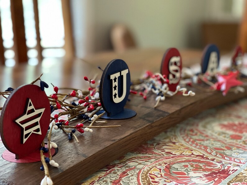 May include: A wooden centrepiece featuring the letters "USA" in red, white and blue. The letters are on individual stands and are surrounded by red berries and white sprigs.