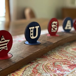 May include: A wooden barrel with five round wooden cutouts painted in red, white and blue. The cutouts spell out "USA" with a star in the centre of the "U" and a star on the right side of the barrel.