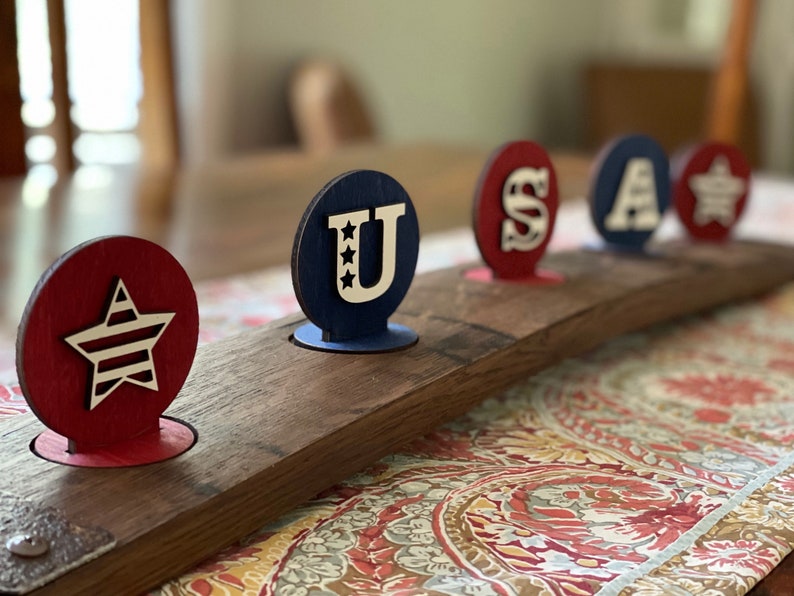 May include: A wooden barrel with five round wooden cutouts painted in red, white and blue. The cutouts spell out "USA" with a star in the centre of the "U" and a star on the right side of the barrel.