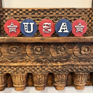 May include: Five wooden circles painted in red, white and blue, spelling out "USA". Each circle features a star with red and white stripes in the centre.