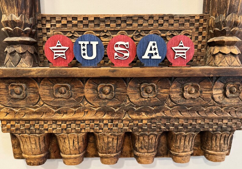 May include: Five wooden circles painted in red, white and blue, spelling out "USA". Each circle features a star with red and white stripes in the centre.