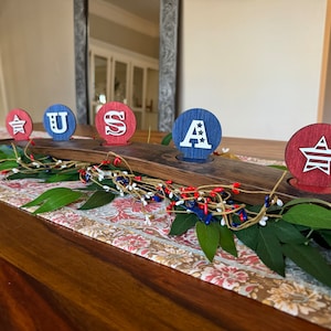 May include: A wooden centrepiece with five round wooden candle holders. The holders are painted red, white, and blue and feature the letters "U.S.A." and a star. The centrepiece is decorated with greenery and red, white, and blue berries.