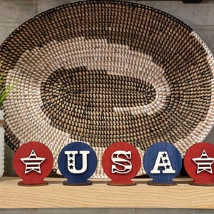May include: Five wooden circles painted in red, white and blue. The circles spell out "USA" with a star on each end. The circles are sitting on a wooden shelf in front of a woven basket.
