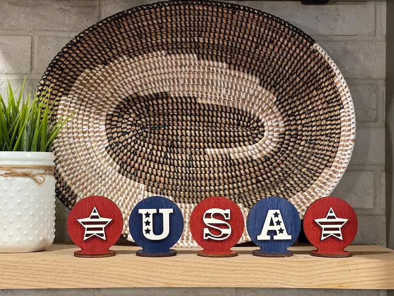 May include: Five wooden circles painted in red, white and blue. The circles spell out "USA" with a star on each end. The circles are sitting on a wooden shelf in front of a woven basket.