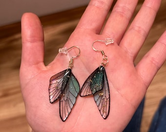 May include: A pair of dangle earrings featuring a layered butterfly design with black wire wings and green and gold glitter accents.