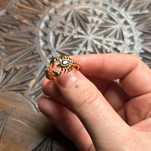 Op de afbeelding: Een goudkleurige ring met een krabontwerp. De krab heeft gedetailleerde klauwen en een getextureerd lichaam, met een witte, ovaalvormige steen in het midden. De ring wordt in een hand gehouden tegen een gebeeldhouwd houten oppervlak.