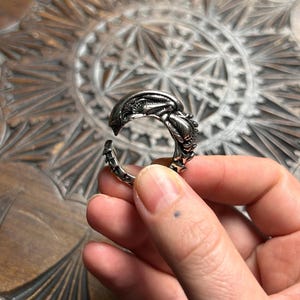 Puede incluir: Un anillo plateado con un diseño detallado de una criatura alienígena. El anillo está abierto en la parte inferior y se sostiene en una mano. El diseño del anillo presenta la cabeza y el cuerpo del alienígena, con detalles intrincados.