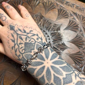 Może przedstawiać: Srebrna bransoletka z łańcuszkiem o unikalnym wzorze węzła. Bransoletka jest na nadgarstku.