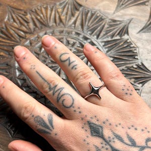 May include: A silver-colored starburst ring is worn on a finger. The hand has several tattoos, including small dots and script. The ring's design features four pointed rays extending from a central point. The background is a carved wooden surface.