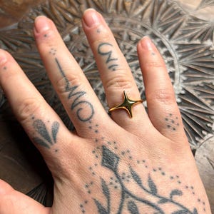 May include: A gold-colored starburst ring. The ring features a four-pointed star design. The ring is worn on a finger. The hand has several tattoos.