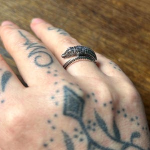 May include: A silver-toned metal ring shaped like a crocodile, featuring detailed scales and a textured surface. The design wraps around the finger, with the crocodile head resting on top and the tail forming the lower part of the band.