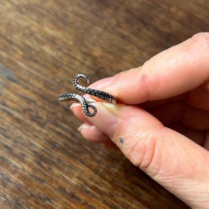 May include: A silver-toned ring shaped like a curled octopus tentacle, featuring a textured surface with small, raised circular suction cup details along the entire length of the band. The metal has a darkened, oxidized finish in the crevices.