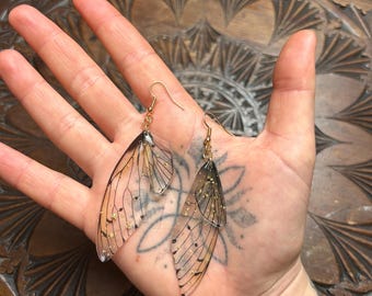 May include: A pair of gold dangle earrings with clear wings and gold glitter accents. The wings are translucent and have a delicate, intricate design.