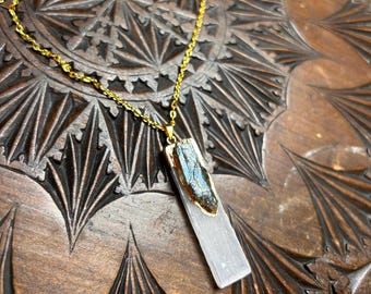 May include: A gold chain necklace with a pendant featuring a clear, rectangular piece of selenite with a dark grey, raw crystal embedded in the center.