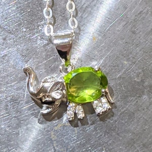 May include: A silver elephant pendant necklace with a green peridot gemstone. The elephant is adorned with small clear cubic zirconia stones.