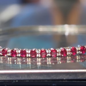Puede incluir: Primer plano de una pulsera de tenis con engaste plateado y numerosas piedras preciosas rojas vibrantes. La pulsera se muestra sobre una superficie reflectante, mostrando los intrincados detalles de la joyería.