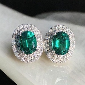 May include: A pair of emerald green oval-shaped earrings with a halo of small diamonds around each stone.