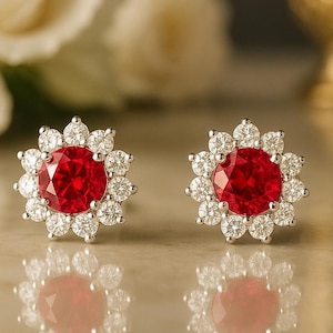 May include: A pair of stud earrings featuring a large, round, vibrant red gemstone surrounded by a halo of smaller, clear gemstones. The earrings are set in a silver-colored metal and reflect light, creating a sparkling effect.