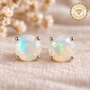 May include: A pair of round, faceted opal stud earrings set in four-prong metal settings. Each opal displays a milky white base with iridescent flashes of green, blue, and orange. A gold-colored seal reads IGI CERTIFIED GEMOLOGIST.