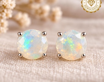 May include: A pair of round, faceted opal stud earrings set in four-prong metal settings. Each opal displays a milky white base with iridescent flashes of green, blue, and orange. A gold-colored seal reads IGI CERTIFIED GEMOLOGIST.