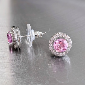 May include: A pair of silver earrings with pink gemstones. The earrings have a round shape and are set with small clear stones around the edges.
