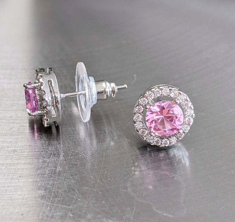 May include: A pair of silver earrings with pink gemstones. The earrings have a round shape and are set with small clear stones around the edges.