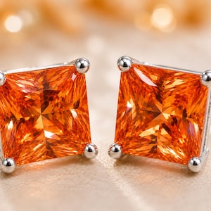 May include: A pair of stud earrings featuring square-cut, vibrant orange gemstones set in polished silver-toned metal. Each earring is secured by four small prongs at the corners, showcasing a faceted surface that reflects light across the stone.