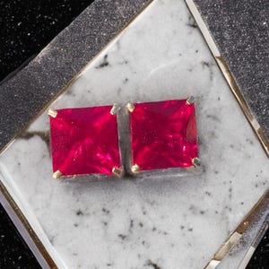 May include: A pair of square-shaped ruby red earrings with gold settings. The earrings are displayed on a white marble surface.