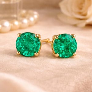 May include: A pair of gold stud earrings featuring large, round, faceted emerald green gemstones. The earrings have a classic four-prong setting. They are displayed on a soft, neutral-toned surface, with pearls and a flower in the background.