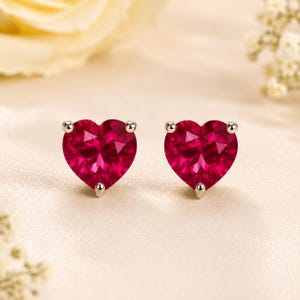 May include: A pair of stud earrings featuring vibrant red, heart-shaped faceted gemstones. Each stone is secured by three small, polished silver-toned metal prongs at the top and bottom points, creating a classic and symmetrical jewelry design.