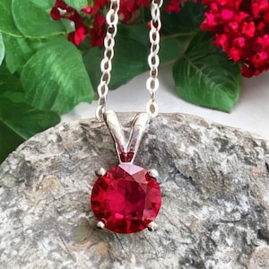 May include: A silver chain necklace with a pendant featuring a round, bright red gemstone.