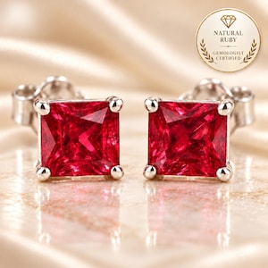 May include: A pair of stud earrings featuring square-cut, deep red rubies set in polished silver-toned metal. Each gemstone is secured by four prongs. A circular gold-toned seal in the upper right corner displays the text Natural Ruby Gemologist Certified.