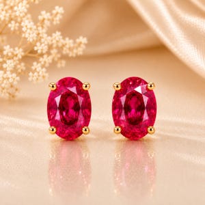 May include: A pair of oval-cut red gemstone stud earrings set in gold-toned metal prongs. Each earring features a faceted, vibrant red stone with a polished finish, secured by four small gold-coloured prongs that hold the gem firmly in place.