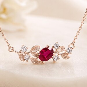 May include: A rose gold necklace featuring a central oval-cut deep red ruby gemstone. The pendant is flanked by textured metal leaves and four small, round, clear white sparkling stones arranged symmetrically on each side of the ruby.