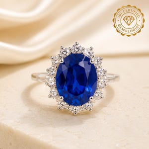 May include: A silver-toned ring featuring a large, oval-cut deep blue center stone surrounded by a halo of small, round, clear stones. The band is thin and metallic. A gold-colored seal in the corner reads IGI CERTIFIED GEMOLOGIST.