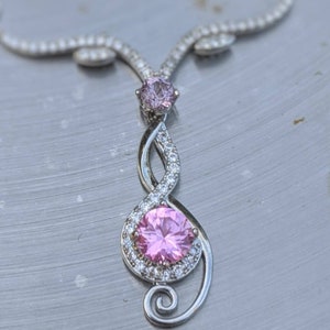 May include: A silver necklace with a pink gemstone pendant. The pendant is shaped like a treble clef and features a smaller pink gemstone at the top.