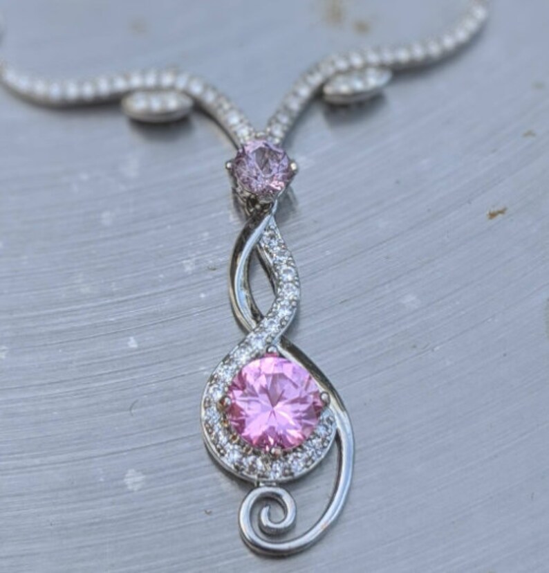 May include: A silver necklace with a pink gemstone pendant. The pendant is shaped like a treble clef and features a smaller pink gemstone at the top.
