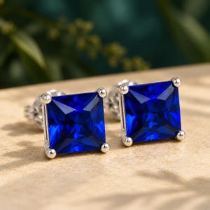 May include: A pair of stud earrings featuring square-cut, deep blue gemstones set in a four-prong silver-toned metal setting. The stones have a faceted surface that reflects light, and the earrings include standard metal backings for secure attachment.