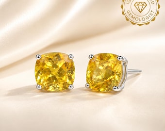 May include: A pair of stud earrings featuring cushion-cut yellow gemstones set in silver-toned metal prongs. Each earring displays a faceted, vibrant yellow stone with a clear, brilliant sparkle. An IGI Certified Gemologist seal is visible.