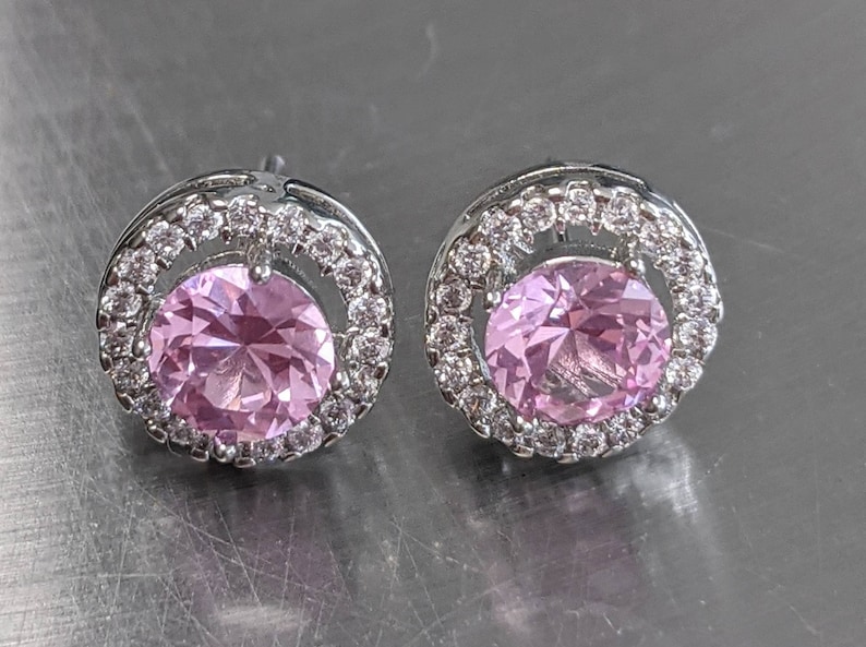 May include: A pair of silver stud earrings with a pink gemstone surrounded by a halo of smaller clear gemstones.