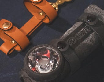 May include: A close-up of a black headlamp with a bright light and a brown leather strap. The strap has gold-coloured hardware. A brown leather key fob with a gold-coloured clasp is also visible.