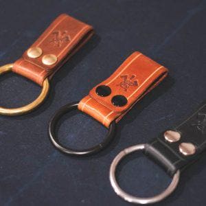 Pode incluir: Três chaveiros de couro com diferentes cores de couro e anéis de metal. Os chaveiros têm um pequeno emblema de metal com um design.